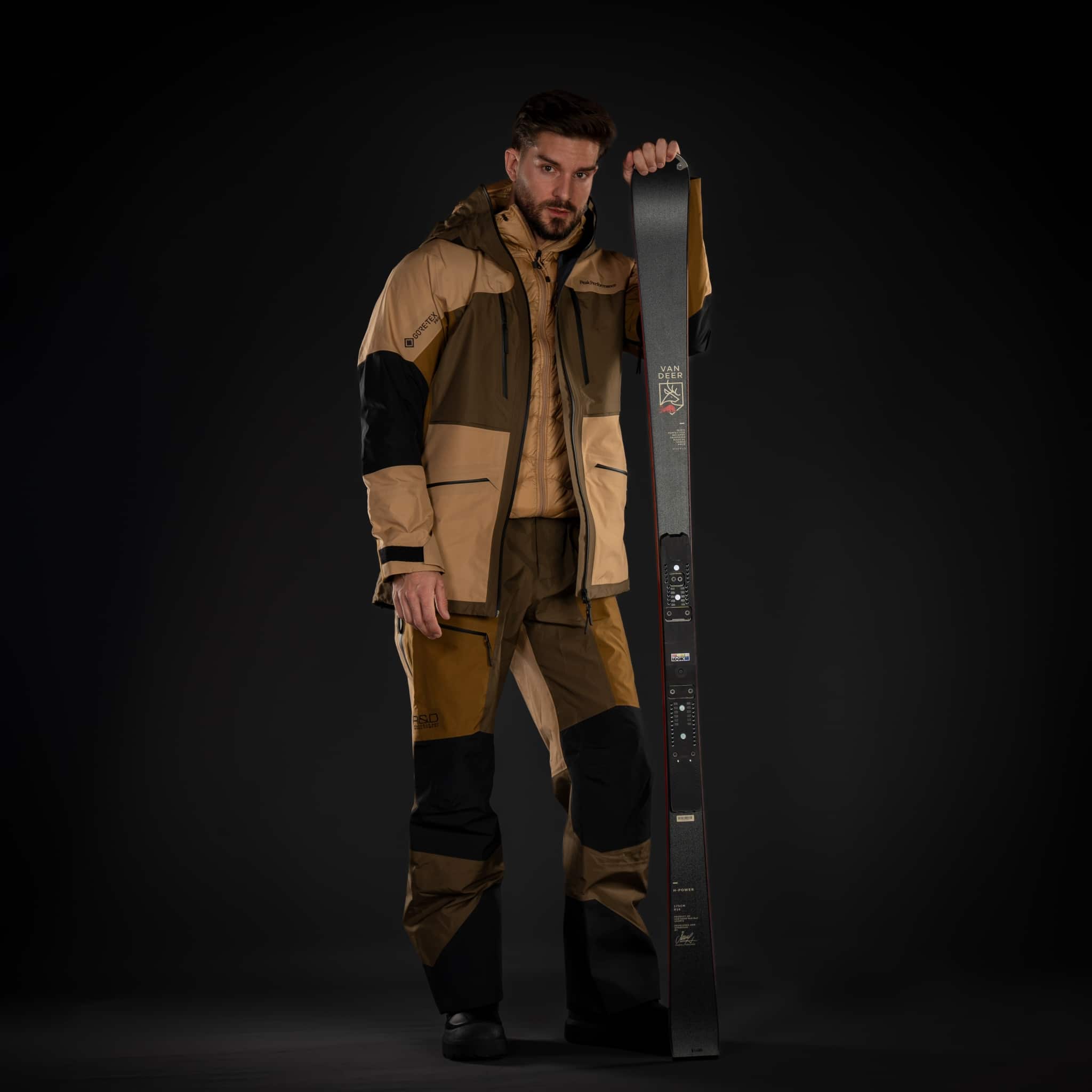 Studioaufnahme: Skifahrer in beige-braunem Skioutfit hält einen Van Deer Ski aufrecht vor schwarzem Hintergrund